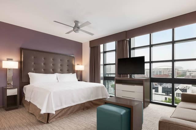 King bed, bedside tables with lamps, couch, TV, dresser, ceiling fan, and large-window outside view