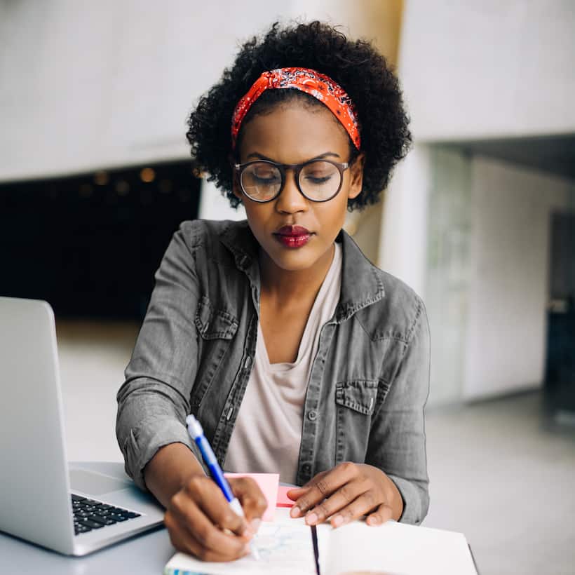 Woman working at a desk with laptop, writing in a notebook